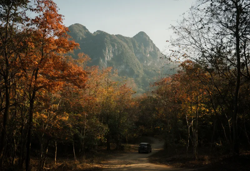 A dirt road through an autumn forest with orange foliage, a small vehicle, and distant mountain peaks (AI-generated)