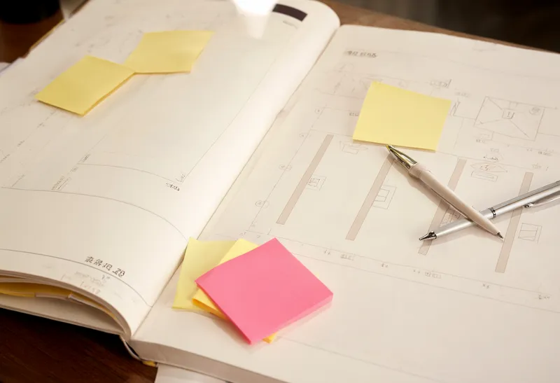 Open design notebook with layout sketches, yellow and pink sticky notes, and two pens arranged on a wooden desk. (AI-generated)