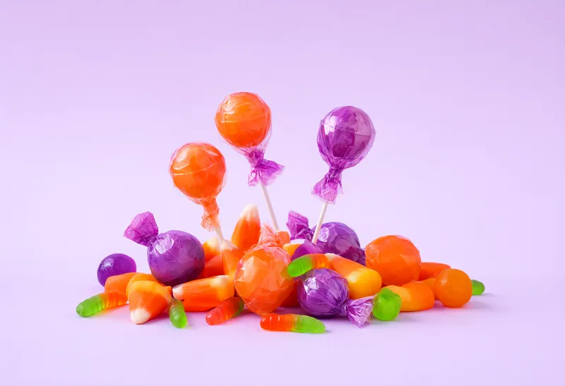 Orange, purple, yellow, and green candies with wrapped lollipops arranged on a light lavender background. (AI-generated)